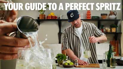 Rico pouring a cocktail into an ice-filled glass while preparing a spritz drink at a kitchen counter, with limes, fresh herbs, and bottles nearby.