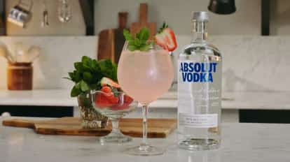 A pink rhubarb spritz cocktail in a stemmed glass, garnished with fresh mint and a strawberry, displayed beside a bottle of Absolut Vodka and fresh strawberries on a kitchen counter.