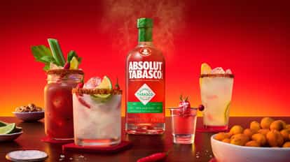 ABSOLUT® TABASCO™ bottle centered on a table, surrounded by a Bloody Mary in a jam jar with celery garnish, a Bloody Mary shot, a spicy lemonade and a spicy vodkarita with chili-salt rim, alongside brunch snacks, set against a vibrant red background.
