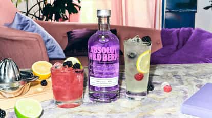 Absolut Wild Berri vodka bottle with two berry cocktails garnished with lemon slices and blackberries on a marble table in a colorful living room setting.