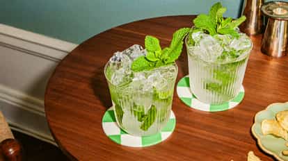 Two iced mint cocktails in ribbed glass tumblers sit on a round wooden table, garnished with fresh mint sprigs and served on green-and-white checkered coasters.