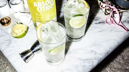 Two Lemon Vodka Soda in highball glasses with ice and lime slices beside an Absolut Citron bottle on a marble table.