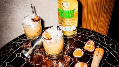Absolut Mango Passion in two rocks glasses with crushed ice and halved passionfruit, bottle and jigger on mosaic table