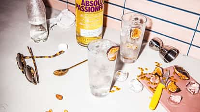 Absolut Passionfruit Fizzy in a highball glass with ice and a passionfruit slice, bottle and bar tools on tiled counter