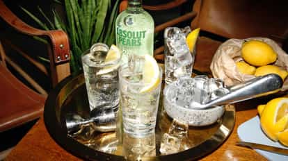 Absolut Pears Lemonade in a tall glass with ice and lemon wedge on a brass tray with bottle behind