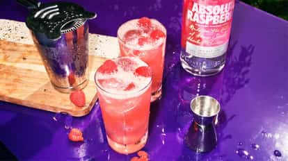 Absolut Raspberri Collins in two Collins glasses with raspberries and ice alongside a bottle and metal shaker and jigger