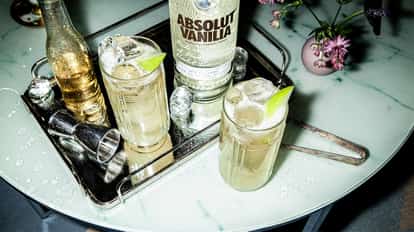 A bottle of Absolut Vanilia vodka with two highball glasses of iced cocktail garnished with lime on a mirrored tray