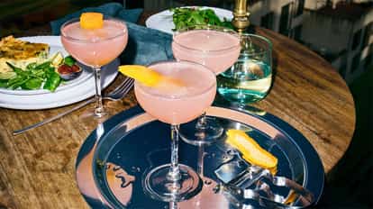 Absolut Wild Berri Cosmo served in three coupe glasses with orange peel garnishes on a metal tray on a wooden table