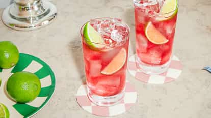 Two glasses of vodka cranberry with ice, lime wedges, and a pink-red color on a marble table.
