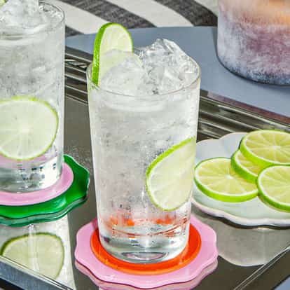 Absolut Lime Soda with ice and lime slices in tall glasses on a mirrored tray with colorful coasters and extra citrus slices.