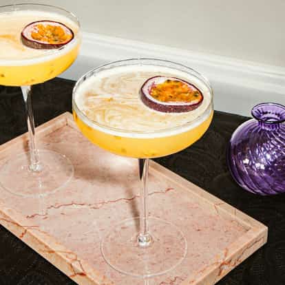 Two passion fruit martinis in coupe glasses with half a passion fruit garnish, served on a pink marble tray.