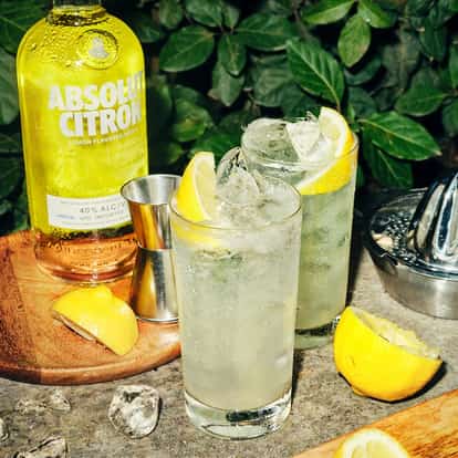 Absolut Citron Lemonade two tall glasses with ice and lemon wedges beside a bottle labeled Citron and a juicer