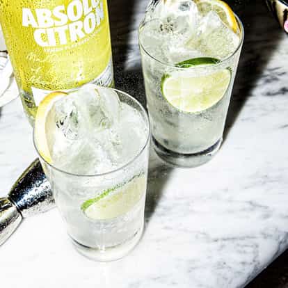 Two Lemon Vodka Soda in highball glasses with ice and lime slices beside an Absolut Citron bottle on a marble table.