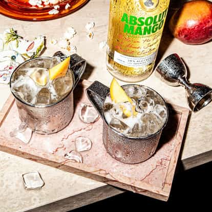 A Moscow Mule variation, the Absolut Mango Mule, served in an Aluminum Mule Mug with lots of ice served on a wooden cutting board with a bottle of Absolut Mango in the background.