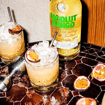 Absolut Mango Passion in two rocks glasses with crushed ice and halved passionfruit, bottle and jigger on mosaic table