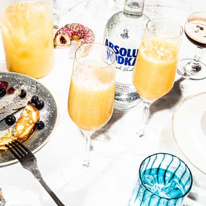 Two mimosas in champagne flutes beside an Absolut vodka bottle and donuts on white background