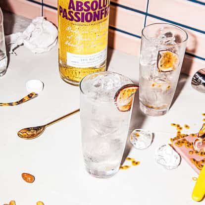 Absolut Passionfruit Fizzy in a highball glass with ice and a passionfruit slice, bottle and bar tools on tiled counter