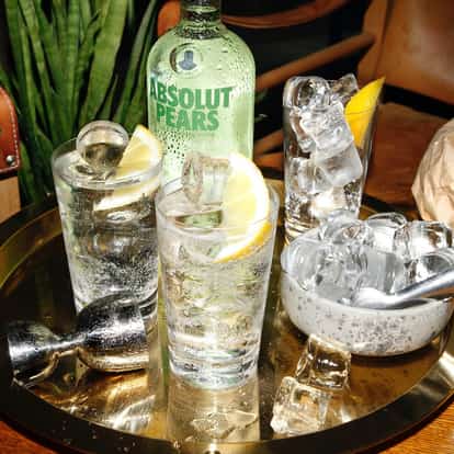 Absolut Pears Lemonade in a tall glass with ice and lemon wedge on a brass tray with bottle behind