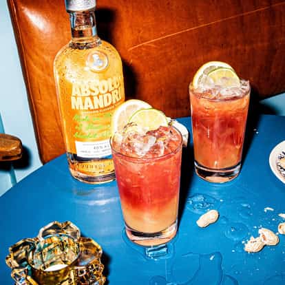 Two punch cocktails in highball glasses with lime wheels beside an Absolut Mandrin bottle on a blue table
