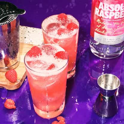 Absolut Raspberri Collins in two Collins glasses with raspberries and ice alongside a bottle and metal shaker and jigger