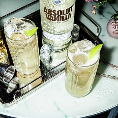 A bottle of Absolut Vanilia vodka with two highball glasses of iced cocktail garnished with lime on a mirrored tray