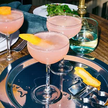 Absolut Wild Berri Cosmo served in three coupe glasses with orange peel garnishes on a metal tray on a wooden table