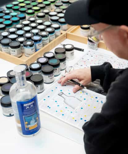 KAWS painting at his studio desk, surrounded by jars of paint and an Absolut bottle