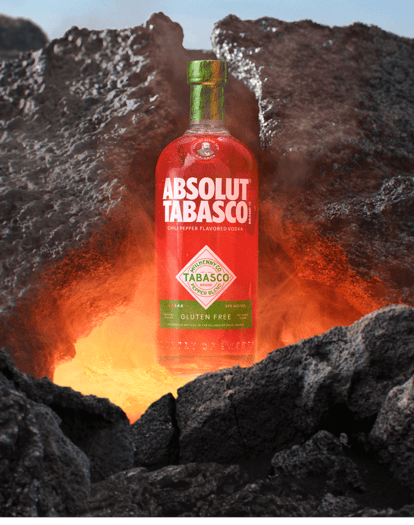 ABSOLUT® TABASCO™ chili pepper flavored vodka bottle emerging from molten lava between dark volcanic rocks, glowing with heat and intensity.