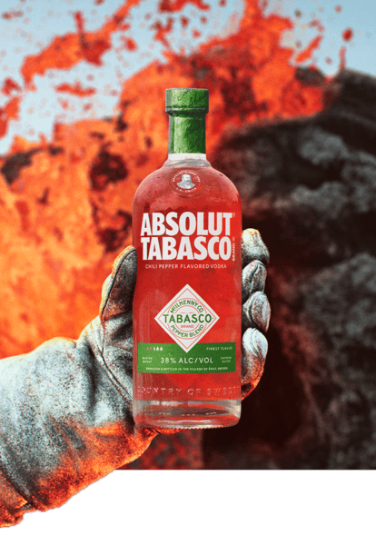 Dramatic shot of a gloved hand holding an ABSOLUT® TABASCO™ vodka bottle in front of erupting lava and dark volcanic rock.