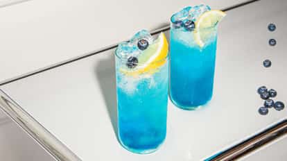 Blueberry Fizz cocktails served in tall, chilled glasses, vibrant blue and packed with ice, garnished with lemon wedges and fresh blueberries.