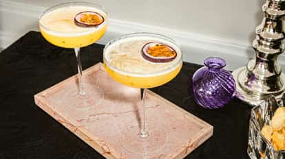 Two passion fruit martinis in coupe glasses with half a passion fruit garnish, served on a pink marble tray.