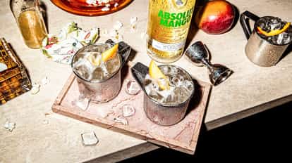 A Moscow Mule variation, the Absolut Mango Mule, served in an Aluminum Mule Mug with lots of ice served on a wooden cutting board with a bottle of Absolut Mango in the background.