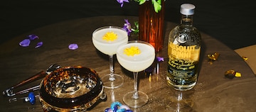 Image of Absolut Hunni bottle presented on a round wooden table compained with 2 coupe glasses of Absolut Hunni lemon drop cocktail.