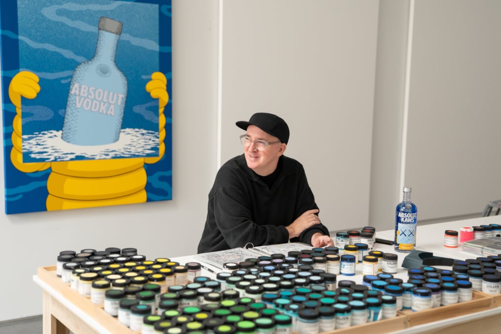 KAWS in his studio with the Absolut KAWS Artist Edition bottle on the table and his commissioned painting of the Absolut Vodka bottle on the wall behind him.