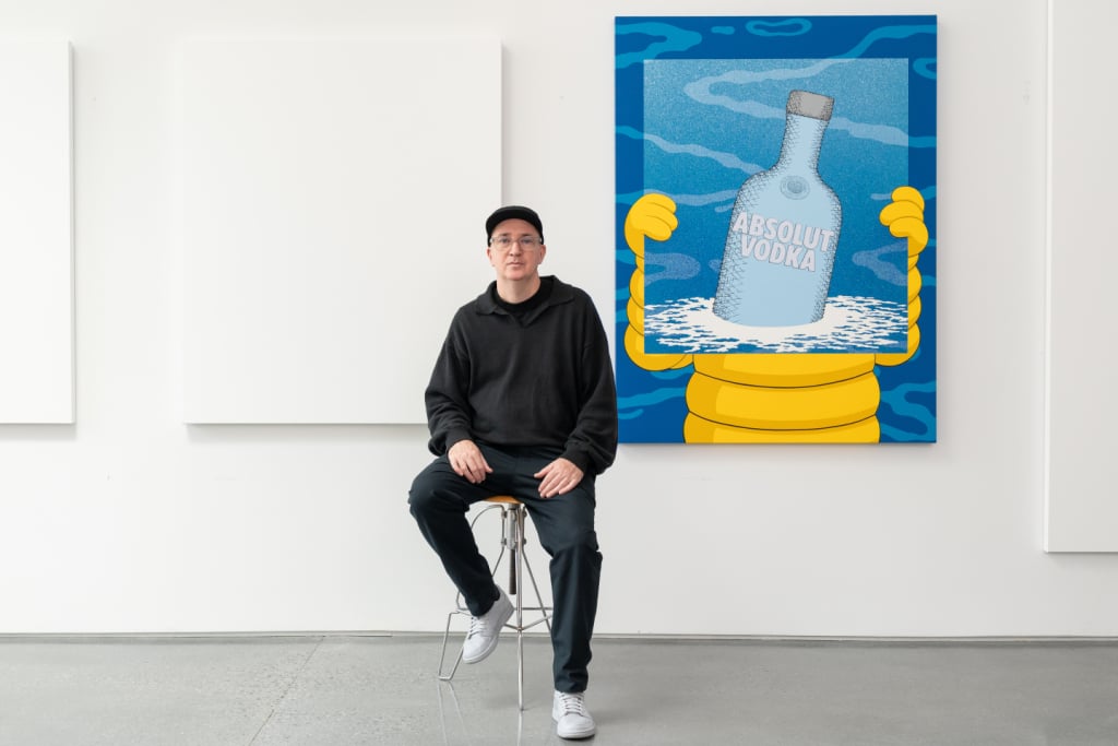 KAWS seated in his studio beside his large-scale commissioned painting of the Absolut Vodka bottle, held by his iconic CHUM character against a blue background.
